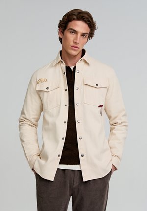 Camisa beige de botones hecha de algodón, con dos bolsillos frontales, que presenta un diseño de sol bordado en marrón y un acento de tela roja cerca del bolsillo.
