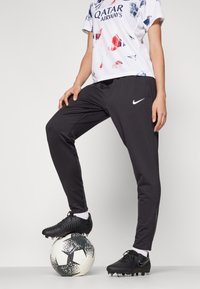 Sorte idrettsbukser med slim fit og hvit Nike-logo, kombinert med en hvit, rød og blå grafisk t-skjorte, iført med svarte fotballsko.