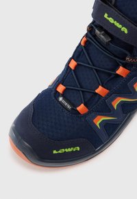 Zapatilla de senderismo Lowa en azul marino con acentos naranjas, etiqueta Gore-Tex, cordones elásticos y correa de velcro, sobre un fondo blanco.