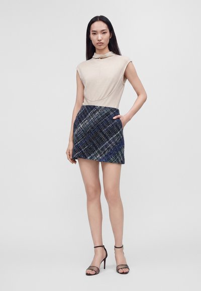 Femme aux longs cheveux foncés portant un haut beige sans manches, une mini-jupe à carreaux bleu marine et des sandales noires à talons hauts, debout devant un fond uni.