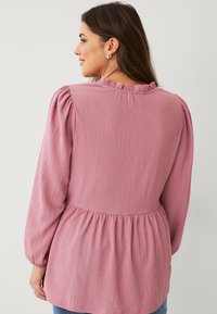 Long-sleeve blouse in light pink textured fabric, featuring a ruffled neckline and a gathered waist for a relaxed fit.