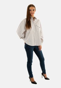 Camisa de botones a rayas en gris claro y blanco, corte oversize, con dos bolsillos frontales, combinada con jeans ajustados de color azul oscuro y tacones negros.