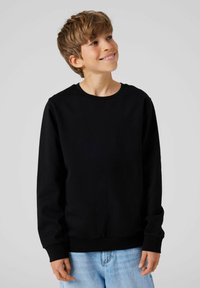 Chico adolescente sonriente con cabello castaño claro, vistiendo un sudadera negra y unos vaqueros de mezclilla azul claro, mirando ligeramente hacia la derecha.