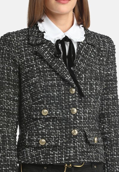 Mustavalkoinen tweed-blazer, jossa on teksturoitu pinta, kaksirivinen muotoilu, kultaiset napitusyksityiskohdat ja valkoinen röyhelökauluspaidat alla.