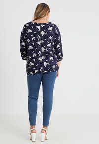 Tummansininen paita, jossa on valkoinen kukkakuosi, rento malli, kolmeeljäsosaa hihat ja takana nappiyksityiskohta, yhdistettynä sinisiin skinny-farkkuihin ja valkoisiin korkokenkiin.