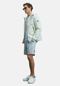 Veste imperméable blanche avec poches zippées, t-shirt bleu clair, shorts gris et baskets blanches avec accents bleus, en pose debout.