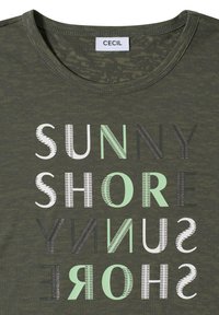 T-shirt vert foncé à col rond avec étiquette "CECIL", affichant le texte stylisé "SUNNY SHORE" répété selon différents motifs blancs, verts et noirs.