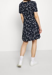 Kvinna som bär en marinblå blommig klänning med korta ärmar och vita sneakers, står mot en slät vit bakgrund, vänd bort.