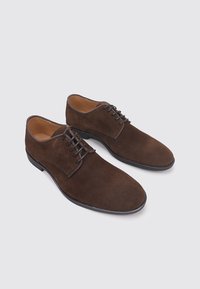 Par de zapatos de vestir para hombre de ante marrón con suelas y cordones negros, exhibidos sobre un fondo gris claro.