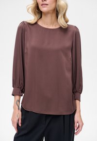 Brązowa bluzka z satyny o okrągłym dekolcie, rękawach trzy-czwarte i marszczonych mankietach. Gładka faktura, luźny krój, w połączeniu z czarnymi spodniami.