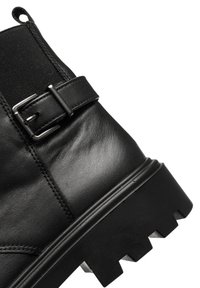 Bottine en cuir noir avec une attache à boucle, des côtés élastiques et une semelle en caoutchouc texturé avec des motifs de traction en zigzag.