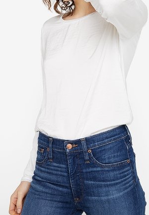 Weißes Longsleeve-Oberteil mit Rundhalsausschnitt, kombiniert mit dunkelblauen Jeans aus Denim mit bronzefarbenen Akzenten und klassischem Fünf-Taschen-Design.