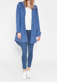 Blauwe waterdichte jas met een capuchon, togglesluitingen, lange mouwen en een gestreepte binnenkant. Gekozen in combinatie met denim jeans en witte sneakers.