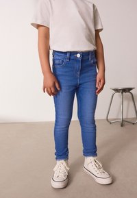 Modré džínsové skinny nohavice s vysokým pásom, obsahujúce predné vrecká a zips, v kombinácii s bielymi vysokými teniskami a svetlým tričkom.