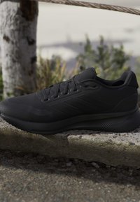 Pantof sport negru cu design cu șireturi pe un prag de piatră în aer liber, lângă un copac și vegetație neclară în fundal.