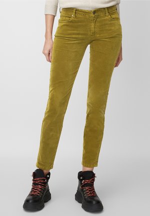 Slim fit mosterdgele corduroy broek gedragen met zwarte wandelschoenen met rode veters, gecombineerd met een crèmekleurige trui, effen grijze achtergrond.