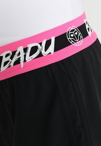 Černé šortky s růžovým elastickým pasem s bílým nápisem "BADU" a grafickým logem. Měkká látka, volný střih, se zřetelnými bočními švy.