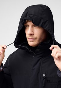 Giacca nera impermeabile con cappuccio regolabile, chiusura con zip e bottoni a pressione, e tasca con zip. Materiale resistente con una finitura testurizzata.