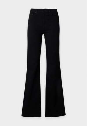 Femme debout portant un haut gris sans manches, un pantalon noir évasé taille haute et des chaussures noires sur un fond clair uni.