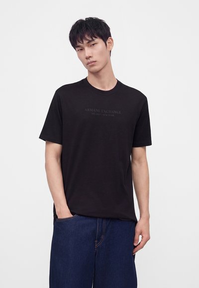 Млад мъж с черна коса, носещ черна тениска Armani Exchange и тъмносини дънки, стои с една ръка в джоба пред бял фон.