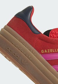 Crvene suede tenisice s mornarsko plavim naglasima, s ružičastim trakama, zlatnim natpisom "GAZELLE" i teksturiranom gumiranom potplatom s utisnutim brendiranjem.