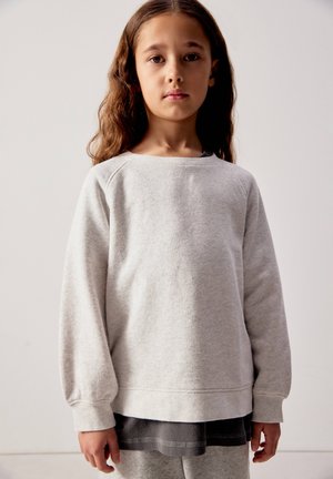 Fille aux longs cheveux bruns portant un sweat-shirt gris clair par-dessus une robe ou une jupe gris foncé, debout devant un fond uni.
