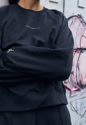 Persoon draagt een zwarte Jordan Sport sweatshirt met gekruiste armen, staand voor een muur met roze en zwarte graffiti.