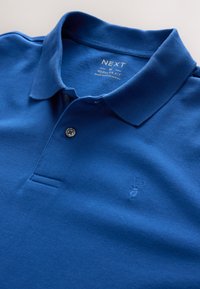 Modré polo tričko strednej veľkosti s pravidelným strihom, dvoma gombíkmi a jemne vyšitým jeleňom na hrudi, vyrobené v Bangladéši.