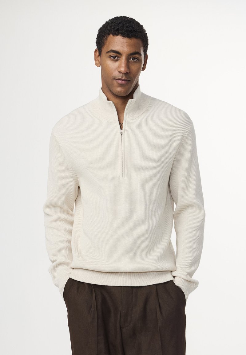 Cremfarbener, gerippter Strickpullover mit einem halben Reißverschluss am Kragen. Merkmale sind lange Ärmel und ein taillierter Saum. Weicher Stoff auf schlichtem Hintergrund.