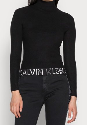 Femme portant un pull à col roulé côtelé noir avec le texte blanc "Calvin Klein" sur l'ourlet, associé à un jean noir avec une bande logo sur le côté.