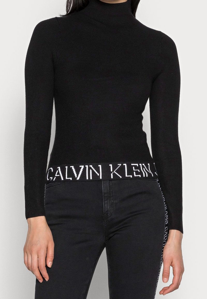 Femme portant un pull à col roulé côtelé noir avec le texte blanc "Calvin Klein" sur l'ourlet, associé à un jean noir avec une bande logo sur le côté.