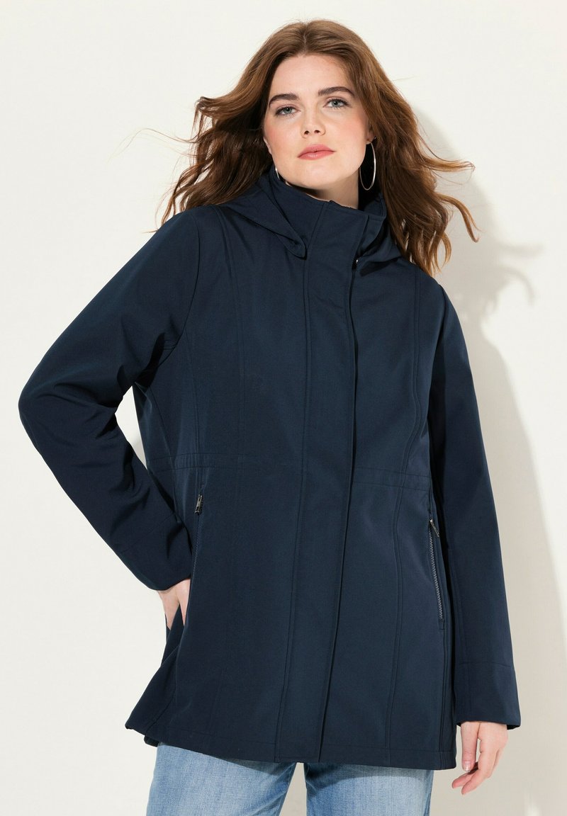 Navy blue pehmeäkuorijakki hupulla, jossa on vetoketjulliset taskut ja istuva muotoilu. Sujuva pinta ja kevyt materiaali.