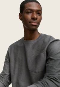 Pilkas ilgomis rankovėmis marškinys su tekstūrinio paviršiaus apdorojimu ir krūtinės kišenėle, pasižymintis apykakle ir paprastu siuvimu, uždedamas ant žmogaus.