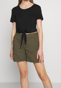 T-shirt noire à nouer devant, manches courtes, associée à un short cargo vert olive avec poches latérales et détails surpiqués.