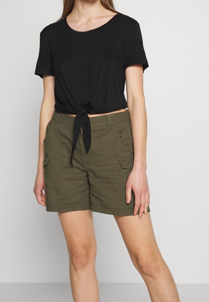 T-shirt noire à nouer devant, manches courtes, associée à un short cargo vert olive avec poches latérales et détails surpiqués.