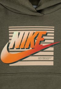 Maslinasto zeleni dukserica s istaknutim narančasto-crnim Nike logom na prugastoj pozadini, s bijelim tekstom ispod. Mekana tkanina.