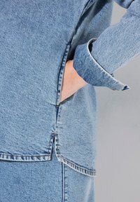 Camisa de mezclilla azul claro con un ajuste ligeramente holgado, con mangas remangadas y bolsillos en las costuras laterales. Detalles de costura visibles a lo largo de los dobladillos.