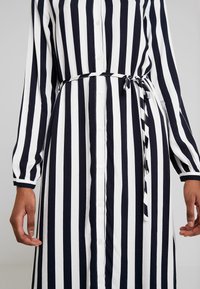 Personne portant une robe boutonnée à rayures verticales noires et blanches, à manches longues et ceinture nouée à la taille.