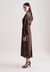 Abito con stampa leopardo in marrone e nero, maniche lunghe con polsini arricciati, vita cintrata, silhouette fluida, abbinato a sandali neri con tacco alto.