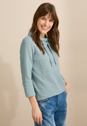 Un pullover texturé bleu clair avec un col rond et un cordon de serrage. Il présente des manches trois-quarts et une coupe décontractée.