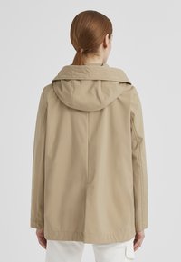 Beige wasserfeste Jacke mit Kapuze, gerade geschnitten, mit glatter Textur und feinen Nahtdetails auf der Rückseite.