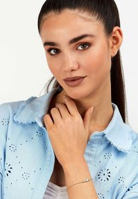 Lyseblå denimskjorte med blomsterbroderi og frayed krave. Guldarmbånd med en dekorativ sten. Model poser med hånden near ansigtet.