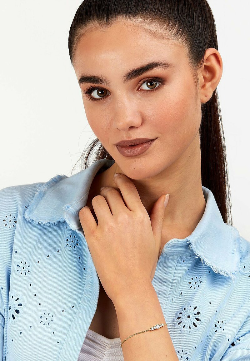 Ljusblå denimskjorta med blommig broderi och fransig krage. Guldarmband med en dekorativ sten. Modellen poserar med handen nära ansiktet.