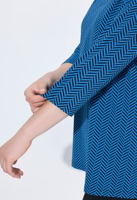 Blusa azul estampada con un diseño de chevrón en negro. Las mangas largas están acampanadas y la tela parece suave y ligera.