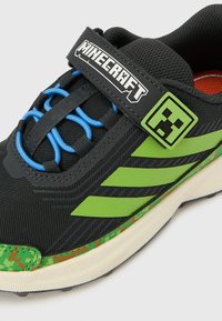 Must ja roheline Minecrafti-teemaline tennisejalanõu siniste elastsete paelte, pikslilise rohelise talladetaili ja logoga rihmal ning küljel.