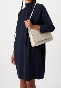 Navy sininen ylisuuri mekko, jossa on pitkät hihat ja korkea kaula-aukko. Vaalea beige käsilaukku, jossa on läppäkiinnitys ja minimalistinen muotoilu, pidetään yhdessä kädessä.