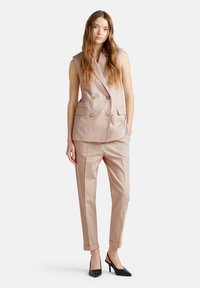 Dubbelgeknoopte mouwloze blazer in lichtroze gestreepte stof, gecombineerd met bijpassende getailleerde broek met opgerolde mouwen en zwarte hakken.