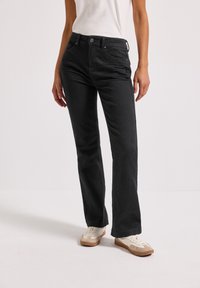 Jean noir à jambe droite en denim, avec une fermeture à boutons, cinq poches et une texture lisse. Portés avec des baskets blanches.