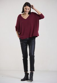 Burgunderfarbener oversized Strickpullover mit V-Ausschnitt, kombiniert mit schwarzen Kunstlederhosen und Ankle Boots, vor einem neutralen Hintergrund.