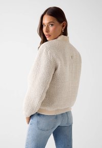 Crèmewitte gebreide trui met geribbelde kraag en zoom, voorzien van een subtiel driehoekig detail aan de achterkant. Gecombineerd met lichtblauwe jeans.